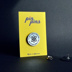 Può includere: Uno spillo argentato con un simbolo di atomo nero e il testo "Think like a proton and stay positive" intorno. Lo spillo è su una carta gialla con il testo "pin pins" in viola e il testo "Made in Lithuania" in basso.