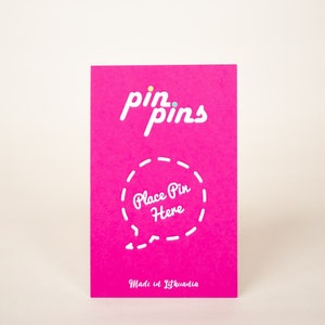 May include: A pink card with the text "pin pins" in white. A white circle with a dashed line border and the text "Place Pin Here" is in the center of the card. The text "Made in Lithuania" is at the bottom of the card.