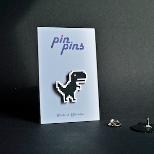 May include: Black pixelated dinosaur pin on a white card with the text "pin.pins" and "Made in Lithuania".