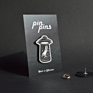 Op de afbeelding: Zwarte en witte emaille pin met een cartoonfiguur dat wordt ontvoerd door een UFO. De pin heeft de tekst "Finally" en "pin pins" op de achterkant. Gemaakt in Litouwen.
