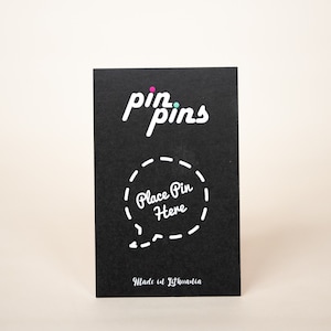 May include: Black card with white text that reads "pin pins" and a dashed circle with the text "Place Pin Here" inside. The card also has the text "Made in Lithuania" at the bottom.