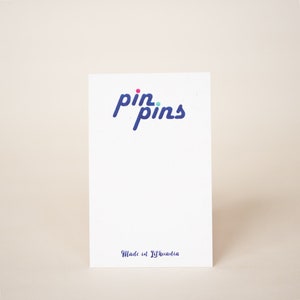 May include: White card with the text "pin.pins" in blue with a pink and green dot above the word "pin". The text "Made in Lithuania" is printed in blue at the bottom of the card.