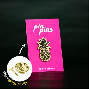May include: Gold pineapple pin with a black outline on a pink background. The pin is on a card that says "pin pins" and "Made in Lithuania". The back of the pin has a butterfly clutch.