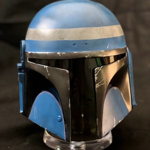 May include: A blue and black Mandalorian helmet with a silver stripe across the top. The helmet has a visor and is displayed on a clear stand. The helmet has a weathered appearance.
