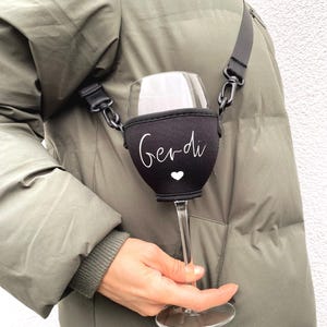 Pode incluir: Um porta-copos de vinho preto com uma alça branca e o nome "Gerdi" escrito nele. O porta-copos está preso ao ombro de uma pessoa.