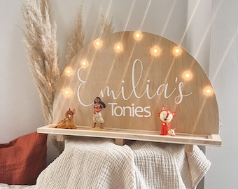 May include: A wooden half-circle sign with a shelf, featuring the name "Emilia's Tonies" in white lettering. The sign has a row of small lights along the top edge. There are three small figurines on the shelf: a deer, a woman, and a fox.