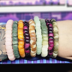 May include: A collection of beaded bracelets in various colors, including pink, green, orange, purple, brown, and white. The bracelets are stacked on a wrist, showcasing the range of hues and textures. The beads appear to be smooth and rounded.