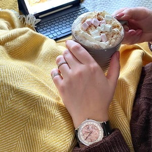 Pu&ograve; includere: Primo piano di un orologio da polso con quadrante in oro rosa e cinturino in pelle nera. L'orologio &egrave; al polso di una persona, che tiene una tazza di cioccolata calda con marshmallow.