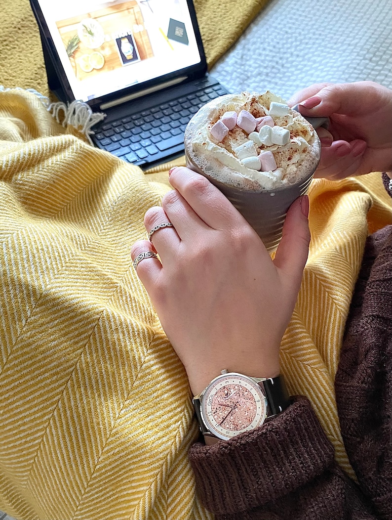 Pu&ograve; includere: Primo piano di un orologio da polso con quadrante in oro rosa e cinturino in pelle nera. L'orologio &egrave; al polso di una persona, che tiene una tazza di cioccolata calda con marshmallow.