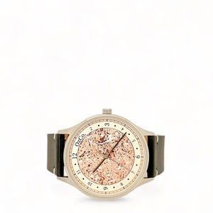 Pu&ograve; includere: Un orologio argentato con un cinturino in pelle marrone. Il quadrante dell'orologio ha uno sfondo color oro rosa con una texture punteggiata e indici delle ore neri. Il quadrante dell'orologio ha anche il testo "12 OzCo."