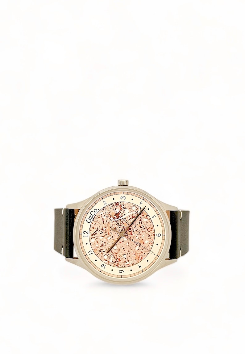 Pu&ograve; includere: Un orologio argentato con un cinturino in pelle marrone. Il quadrante dell'orologio ha uno sfondo color oro rosa con una texture punteggiata e indici delle ore neri. Il quadrante dell'orologio ha anche il testo "12 OzCo."