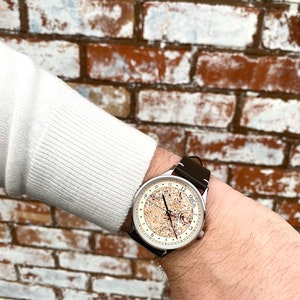 Pu&ograve; includere: Un orologio da polso argentato con quadrante bianco e cinturino in pelle nera. Il quadrante dell'orologio ha un design unico con un motivo marrone e bianco. Il quadrante dell'orologio ha anche indici orari in numeri romani neri e lancette nere.