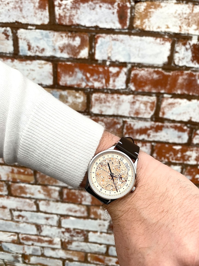 Pu&ograve; includere: Un orologio da polso argentato con quadrante bianco e cinturino in pelle nera. Il quadrante dell'orologio ha un design unico con un motivo marrone e bianco. Il quadrante dell'orologio ha anche indici orari in numeri romani neri e lancette nere.