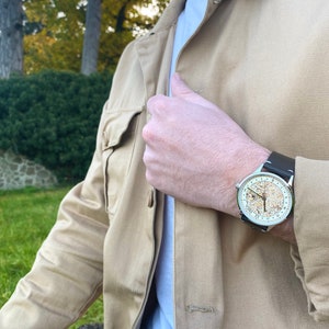 Pu&ograve; includere: Un uomo indossa una giacca beige e una t-shirt bianca con un orologio d'argento con un cinturino in pelle marrone. Il quadrante dell'orologio ha uno sfondo dorato con numeri neri e un anello esterno bianco.