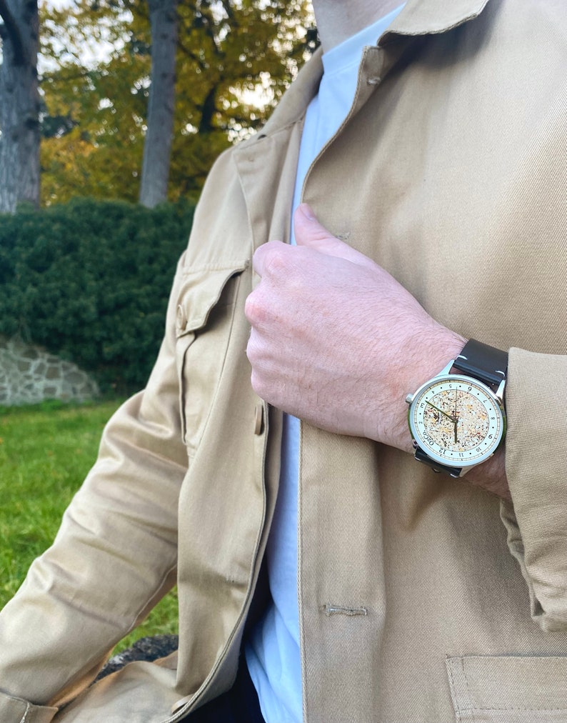 Pu&ograve; includere: Un uomo indossa una giacca beige e una t-shirt bianca con un orologio d'argento con un cinturino in pelle marrone. Il quadrante dell'orologio ha uno sfondo dorato con numeri neri e un anello esterno bianco.