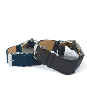 Pu&ograve; includere: Due orologi da polso con cinturini in pelle nera. L'orologio a sinistra ha un cinturino blu e l'orologio a destra ha un cinturino marrone. Entrambi gli orologi hanno casse argentate e l'orologio a destra ha la scritta "Ozco All Stainless Steel Japan Movement" sul retro.