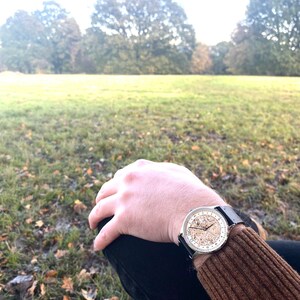 Pu&ograve; includere: Un orologio d'argento con un cinturino in pelle nera e un quadrante bianco con segni neri. L'orologio &egrave; al polso di una persona, e lo sfondo &egrave; un campo erboso con alberi in lontananza.
