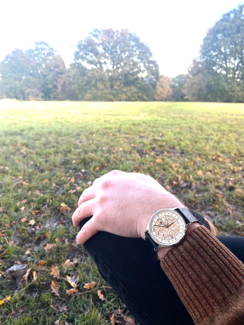 Pu&ograve; includere: Un orologio d'argento con un cinturino in pelle nera e un quadrante bianco con segni neri. L'orologio &egrave; al polso di una persona, e lo sfondo &egrave; un campo erboso con alberi in lontananza.