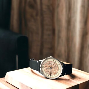 Pu&ograve; includere: Un orologio da polso argentato con un quadrante in sughero e un cinturino in pelle nera. L'orologio &egrave; appoggiato su una superficie di legno.