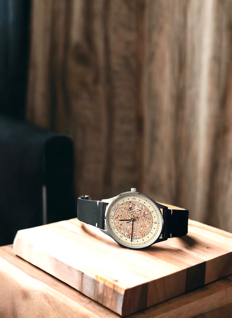 Pu&ograve; includere: Un orologio da polso argentato con un quadrante in sughero e un cinturino in pelle nera. L'orologio &egrave; appoggiato su una superficie di legno.