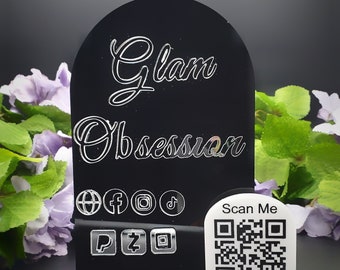 May include: Black acrylic sign with the text "Glam Obsession" in a white cursive font. The sign has a rounded top and a clear base. The sign also includes social media icons and a QR code with the text "Scan Me". The base of the sign has the text "KUSTOMIZED BY COYERA".