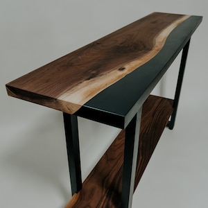 May include: A walnut wood console table with a black epoxy resin inlay and a lower shelf. The table has a modern design with black metal legs.