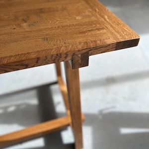 Könnte beinhalten: Nahaufnahme eines Holztisches mit natürlicher Oberfläche. Der Tisch hat eine rechteckige Platte mit sichtbarer Holzmaserung und eine stabile Bein-Struktur. Das Holz hat eine warme, hellbraune Farbe.