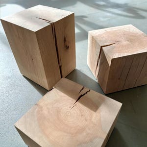 May include: Three light-coloured wooden cubes with visible grain and cracks. The cubes are arranged at different angles, showcasing their natural wood texture. The lighting highlights the details of the wood.