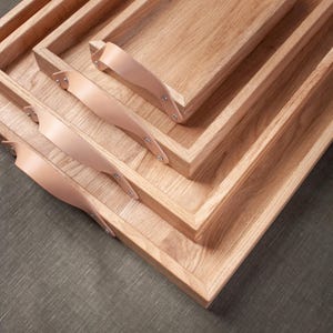 以下が含まれることがあります： 革の取っ手が付いた木製のトレイが4つ、積み重ねられています。トレイは薄い茶色の木でできており、自然な仕上げになっています。革の取っ手は薄い茶色で、トレイの側面に取り付けられています。