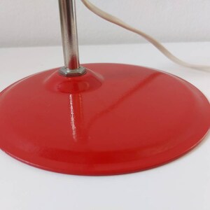 K&ouml;nnte beinhalten: Nahaufnahme eines roten, runden Lampenfu&szlig;es mit silbernem Stiel und wei&szlig;em Stromkabel. Der Sockel hat eine einfarbige, gl&auml;nzende rote Farbe. Die Lampe scheint ein Vintage- oder Retro-Design zu sein.