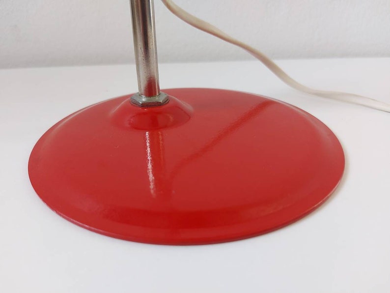 K&ouml;nnte beinhalten: Nahaufnahme eines roten, runden Lampenfu&szlig;es mit silbernem Stiel und wei&szlig;em Stromkabel. Der Sockel hat eine einfarbige, gl&auml;nzende rote Farbe. Die Lampe scheint ein Vintage- oder Retro-Design zu sein.