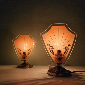 Może przedstawiać: Dwie zabytkowe lampy stołowe w stylu Art Deco. Każda lampa ma klosz w kształcie tarczy z brzoskwiniowym środkiem i czarnymi dekoracyjnymi akcentami. Lampy mają mosiężną podstawę i czerwony przewód zasilający. Lampy są zapalone.