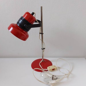 K&ouml;nnte beinhalten: Eine rote und schwarze Vintage-Schreibtischlampe mit Chromstiel und -fu&szlig;. Die Lampe hat einen runden Fu&szlig;, einen zylindrischen Stiel und einen bauchigen Lampenkopf. Die Gl&uuml;hbirne ist sichtbar und die Lampe ist ausgeschaltet.
