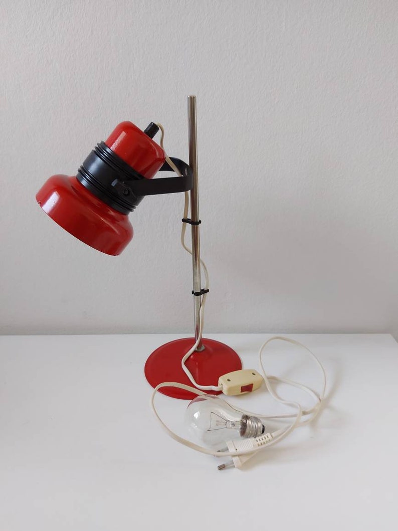 K&ouml;nnte beinhalten: Eine rote und schwarze Vintage-Schreibtischlampe mit Chromstiel und -fu&szlig;. Die Lampe hat einen runden Fu&szlig;, einen zylindrischen Stiel und einen bauchigen Lampenkopf. Die Gl&uuml;hbirne ist sichtbar und die Lampe ist ausgeschaltet.
