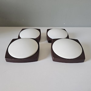 Op de afbeelding: Een set van vier vierkante knopen, elk voorzien van een donkerbruin frame met een glad, verhoogd wit cirkelvormig midden. De knopen zijn gerangschikt in een twee-bij-twee raster en tonen een strak, geometrisch ontwerp met contrasterende kleuren.