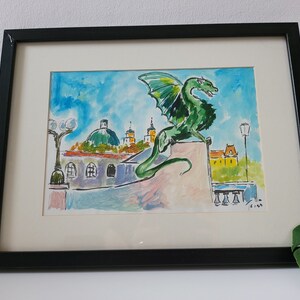 Puede incluir: Una pintura al agua enmarcada de un drag&oacute;n verde posado en un edificio con un cielo azul y nubes blancas en el fondo. El drag&oacute;n est&aacute; mirando hacia la derecha y tiene una cola larga. El edificio es de color marr&oacute;n claro con un techo amarillo. Hay dos farolas a cada lado del drag&oacute;n.