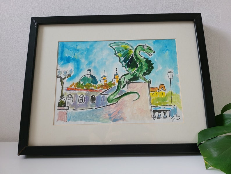 Puede incluir: Una pintura al agua enmarcada de un drag&oacute;n verde posado en un edificio con un cielo azul y nubes blancas en el fondo. El drag&oacute;n est&aacute; mirando hacia la derecha y tiene una cola larga. El edificio es de color marr&oacute;n claro con un techo amarillo. Hay dos farolas a cada lado del drag&oacute;n.