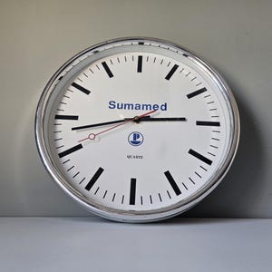 Könnte beinhalten: Eine runde, weiße Quarzuhr mit einem silberfarbenen Metallrahmen. Das Zifferblatt hat schwarze Stundenmarkierungen und Zeiger, mit dem Wort "Sumamed" in Blau. Die Uhr hat auch ein kleines Logo und das Wort "QUARTZ" auf dem Zifferblatt.