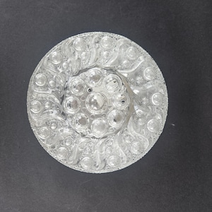 May include: A clear glass ceiling light fixture with a circular design. The fixture has a central cluster of small, round lights surrounded by a decorative pattern of swirls and bubbles. The light is on a black background.