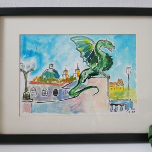 Puede incluir: Pintura acuarela de un drag&oacute;n verde posado en un edificio con una ciudad en el fondo. El drag&oacute;n tiene una cola larga y alas. La ciudad incluye edificios con c&uacute;pulas y torres. La pintura est&aacute; enmarcada en negro.