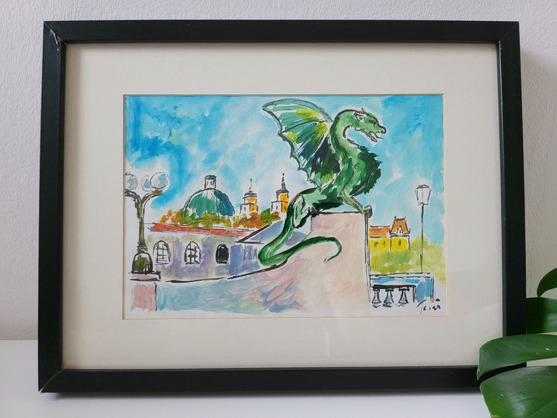 Puede incluir: Pintura acuarela de un drag&oacute;n verde posado en un edificio con una ciudad en el fondo. El drag&oacute;n tiene una cola larga y alas. La ciudad incluye edificios con c&uacute;pulas y torres. La pintura est&aacute; enmarcada en negro.