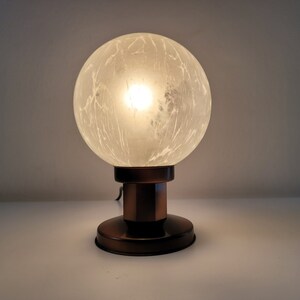 Könnte beinhalten: Eine Tischlampe mit einem mattierten Glaskugelschirm und einem Kupferfuß. Die Lampe ist eingeschaltet und das Licht scheint durch die Kugel.
