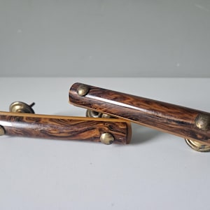 Può includere: Due pomelli vintage per mobili con un motivo a venature del legno nei toni del marrone e del beige. Ogni pomello ha una forma cilindrica con ferramenta color ottone. I pomelli sono progettati per una facile installazione.