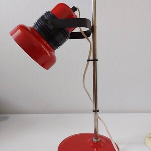 K&ouml;nnte beinhalten: Rote Schreibtischlampe mit schwarzem Akzentring und silberner Stange. Die Lampe hat einen runden Sockel und einen gerichteten Kopf. Das Kabel ist wei&szlig;. Die Lampe steht auf einer wei&szlig;en Oberfl&auml;che.