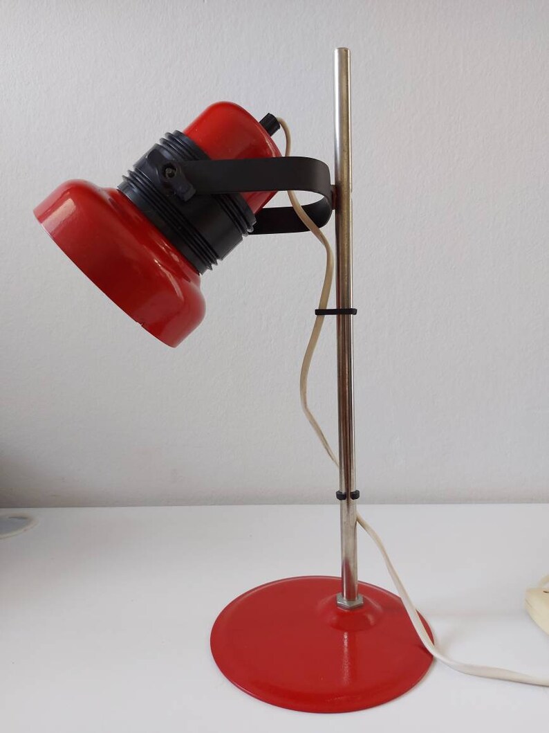 K&ouml;nnte beinhalten: Rote Schreibtischlampe mit schwarzem Akzentring und silberner Stange. Die Lampe hat einen runden Sockel und einen gerichteten Kopf. Das Kabel ist wei&szlig;. Die Lampe steht auf einer wei&szlig;en Oberfl&auml;che.
