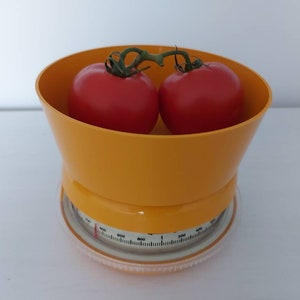 Pode incluir: Uma balança de cozinha amarela com dois tomates vermelhos na tigela. A balança tem um mostrador branco com marcas pretas.