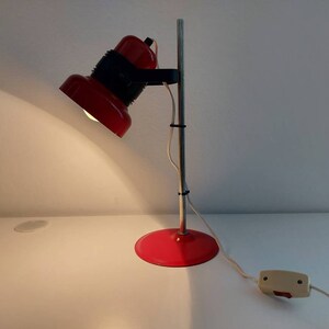 K&ouml;nnte beinhalten: Eine rote Schreibtischlampe mit rundem Fu&szlig; und verstellbarem Kopf. Die Lampe hat einen silbernen Pfosten und ein schwarzes Gelenk, das den Kopf mit dem Pfosten verbindet. Die Lampe ist an eine Steckdose angeschlossen und eingeschaltet, wodurch die darunter liegende Oberfl&auml;che beleuchtet wird.