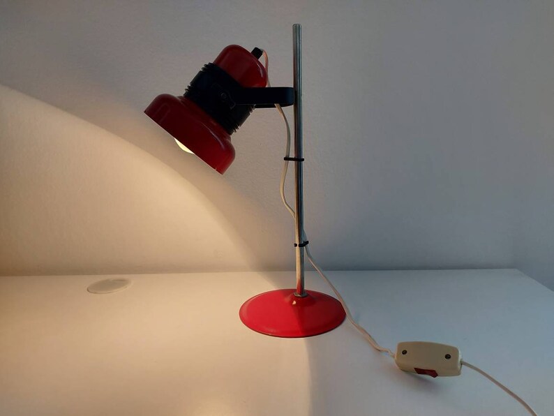 K&ouml;nnte beinhalten: Eine rote Schreibtischlampe mit rundem Fu&szlig; und verstellbarem Kopf. Die Lampe hat einen silbernen Pfosten und ein schwarzes Gelenk, das den Kopf mit dem Pfosten verbindet. Die Lampe ist an eine Steckdose angeschlossen und eingeschaltet, wodurch die darunter liegende Oberfl&auml;che beleuchtet wird.