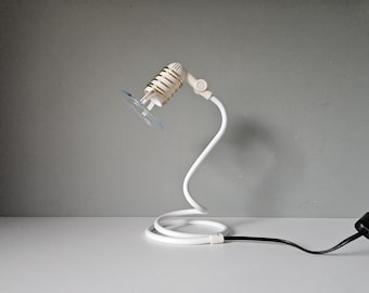 May include: A white, flexible desk lamp with a coiled base and a small, clear suction cup. The lamp head has a cylindrical design with a gold-coloured coil. A black power cord extends from the base.