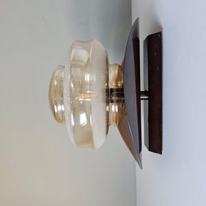 Puede incluir: Aplique de pared vintage con una pantalla de cristal ámbar esmerilado con un diseño acanalado. El aplique está montado sobre una base de madera marrón oscuro con un acento metálico. La lámpara está diseñada para una sola bombilla.